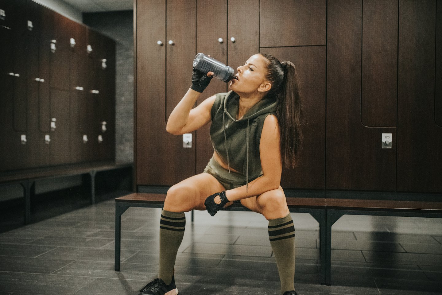 Woman in workout gear drinks water on a gym bench.
